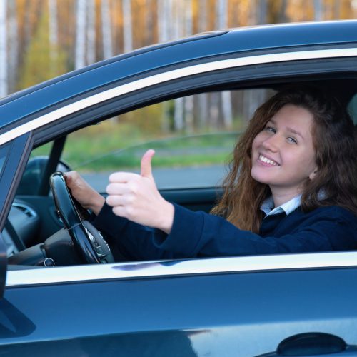 Portrait of happy positive girl, young woman driver is sitting in her car, new automobile, enjoying driving, having fun, laugh. Joyful lady in auto showing thumb up, like gesture